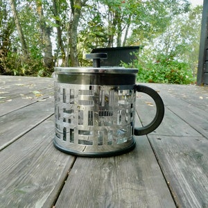 May include: A stainless steel French press coffee maker with a black handle and lid. The body features a decorative cut-out design. The press is sitting on a weathered wooden deck.