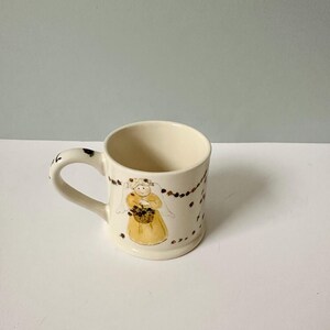 K&ouml;nnte beinhalten: Eine wei&szlig;e Keramiktasse mit Henkel. Die Tasse hat eine braune und gelbe Illustration eines Engels, der einen Blumenkorb h&auml;lt. Der Engel tr&auml;gt einen wei&szlig;en Schleier und ein gelbes Kleid. Der Hintergrund der Tasse ist mit einer braunen und wei&szlig;en Girlande verziert.