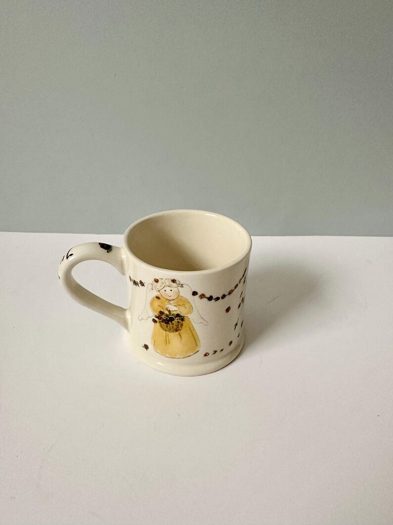 K&ouml;nnte beinhalten: Eine wei&szlig;e Keramiktasse mit Henkel. Die Tasse hat eine braune und gelbe Illustration eines Engels, der einen Blumenkorb h&auml;lt. Der Engel tr&auml;gt einen wei&szlig;en Schleier und ein gelbes Kleid. Der Hintergrund der Tasse ist mit einer braunen und wei&szlig;en Girlande verziert.