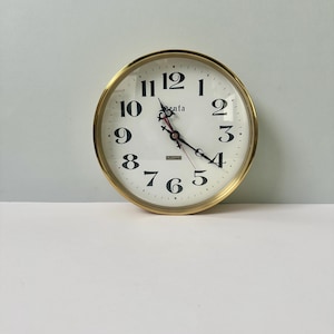 May include: A round wall clock with a metallic gold frame and a white face. The clock features black Arabic numerals from 1 to 12, black ornate hands, a red second hand, and the text "Senta" and "QUARTZ" printed in black on the center dial.