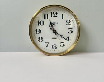 Puede incluir: Un reloj de pared redondo con marco de metal dorado y esfera blanca. El reloj presenta números arábigos negros del 1 al 12, manecillas negras ornamentadas, un segundero rojo y el texto "Senta" y "QUARTZ" impresos en negro en el centro.
