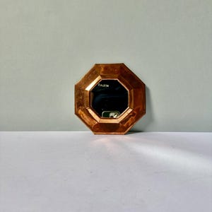 May include: An octagonal mirror featuring a polished copper frame with a faceted, beveled edge design. The reflective surface shows a dark interior reflection with a small white rectangular object visible in the center of the glass.