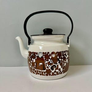 May include: White enamel teapot with a black handle, lid, and rim. The teapot features a brown and white medieval-themed design around the base. The spout is white, and the handle arches over the top.