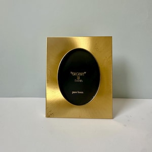 May include: A gold-toned oval picture frame made of pure brass. The frame has a slightly distressed finish and features the text "Gloria" and "Japan" in a smaller font.