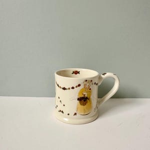 K&ouml;nnte beinhalten: Eine wei&szlig;e Keramiktasse mit einem Blumendesign und einem Bild einer Frau in einem gelben Kleid, die einen Blumenkorb h&auml;lt. Die Tasse hat einen Henkel auf der rechten Seite.