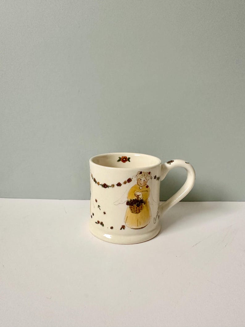 K&ouml;nnte beinhalten: Eine wei&szlig;e Keramiktasse mit einem Blumendesign und einem Bild einer Frau in einem gelben Kleid, die einen Blumenkorb h&auml;lt. Die Tasse hat einen Henkel auf der rechten Seite.