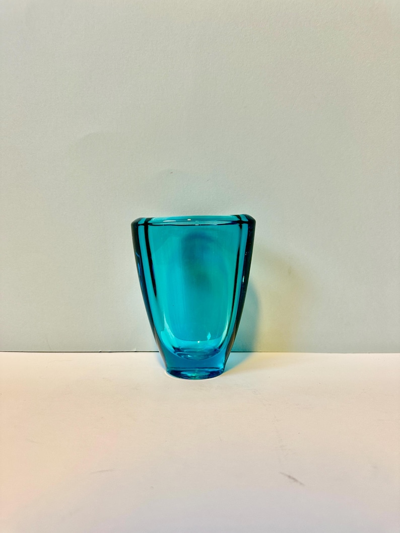 May include: A small, teal blue glass vase with a rounded, slightly flared opening and a flat bottom.