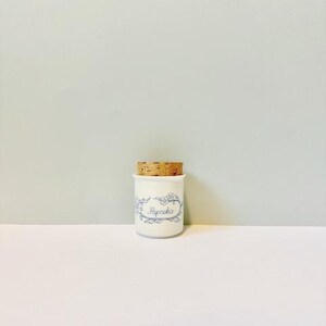 Op de afbeelding: Een witte keramische kruidenpot met een kurken deksel. De pot is versierd met blauwe bloemmotieven en het woord "Paprika" in script. Een keukenartikel.