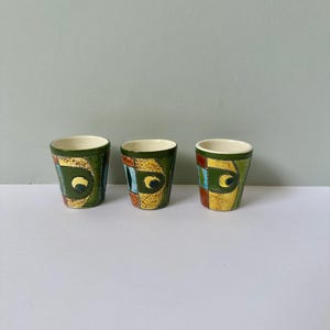 Könnte beinhalten: Drei kleine Keramikbecher mit einem geometrischen Design in Grün, Gelb, Blau und Rot. Die Becher haben ein weißes Innenleben.
