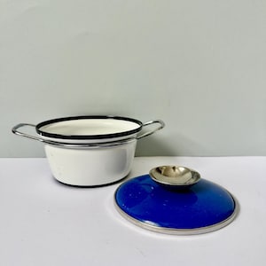 Op de afbeelding: Een kleine, witte emaille pot met een zwarte rand en zilveren handvatten. Een los deksel in kobaltblauw met een zilveren rand en een zilveren knop ligt naast de pot. Een klassiek keukenartikel.