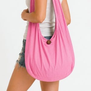 May include: A bright pink hobo-style shoulder bag made of soft fabric, featuring a single wide strap and a prominent round wooden button closure at the center. The bag has a slouchy, unstructured silhouette that hangs comfortably over the shoulder.