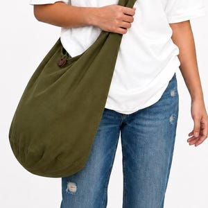 May include: An olive green hobo-style crossbody bag featuring a wide shoulder strap and a single dark brown circular button closure detail. The bag is constructed from a soft, solid-colored fabric and hangs at the hip with a rounded base shape.