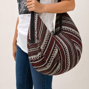 May include: A hobo-style shoulder bag featuring a woven fabric with a repeating pattern of dark red, black, and white stripes, accented by zigzag lines. The bag has a wide, solid black fabric strap that extends from the top opening.
