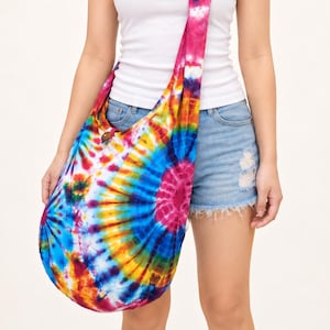 Op de afbeelding: Een soepele hobo-schoudertas met een levendig tie-dye patroon in tinten blauw, geel, oranje en magenta. De tas heeft een brede, geïntegreerde riem die over het lichaam rust en een cirkelvormig zonnestraalontwerp op de stof laat zien.