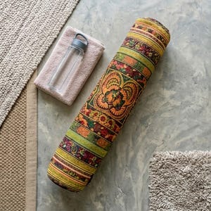 May include: A cylindrical yoga bolster featuring a vibrant, multi-colored woven pattern with shades of orange, green, and yellow, accompanied by a clear plastic water bottle with a grey lid resting on a folded light pink towel.