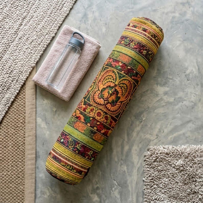 May include: A cylindrical yoga bolster featuring a vibrant, multi-colored woven pattern with shades of orange, green, and yellow, accompanied by a clear plastic water bottle with a grey lid resting on a folded light pink towel.