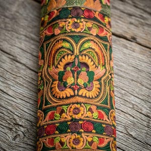May include: A cylindrical textile object featuring dense, multi-colored embroidery in shades of green, orange, yellow, and purple. The surface displays intricate floral and geometric patterns with swirling motifs and symmetrical petal designs.
