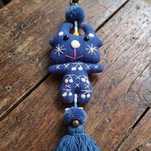 May include: A blue fabric keychain featuring a stylized animal figure with embroidered white star and cross patterns. The figure has black bead eyes, a white nose, and a pink mouth. It is accented with two blue pom-poms, beads, and a long blue tassel.