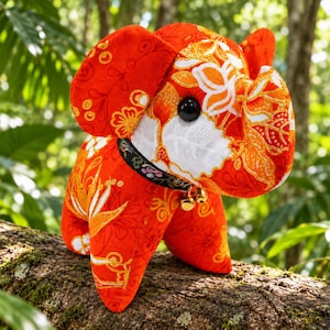 May include: A stuffed elephant toy crafted from vibrant orange fabric featuring a white and gold floral pattern. The elephant has large ears, a trunk, and a black plastic eye. A black collar with a floral design and two small gold bells is worn.