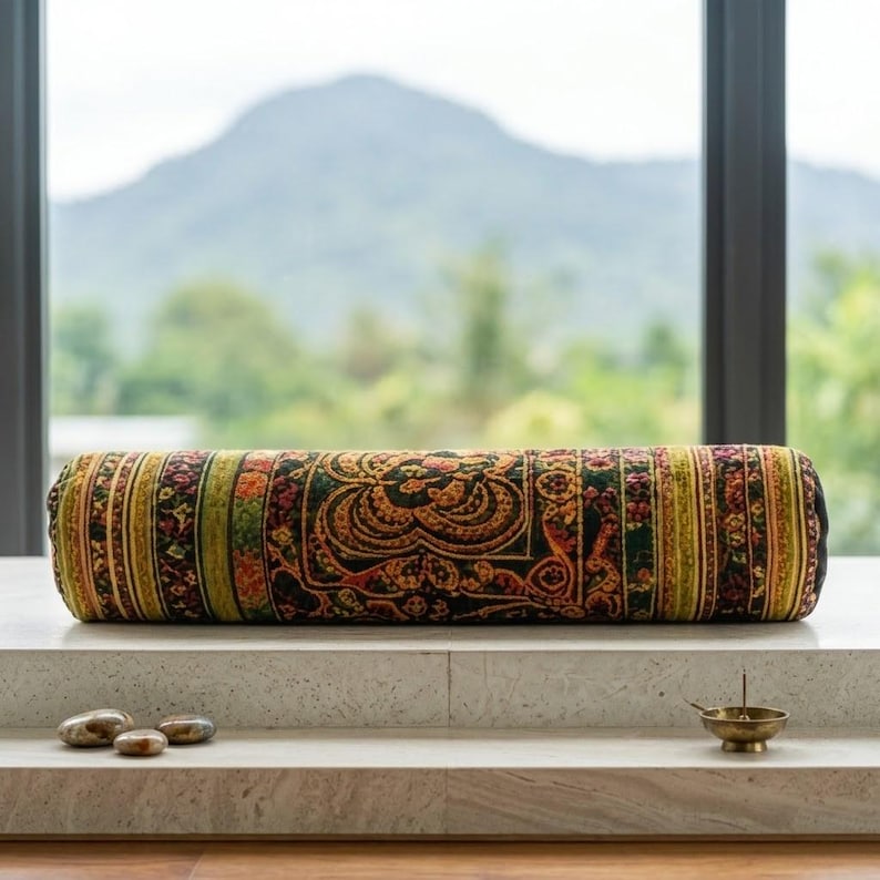 May include: A cylindrical bolster pillow featuring a dark green, orange, and yellow patterned fabric with intricate floral and geometric motifs. The pillow is positioned horizontally, accompanied by three small smooth stones and a brass incense burner.