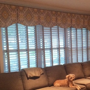 May include: A living room with a brown sectional sofa, a small dog sitting on the sofa, and a window with white blinds and a patterned valance.