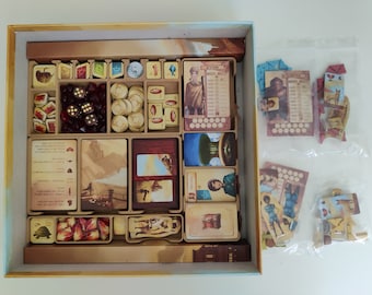 May include: A wooden game organizer with compartments for storing game pieces, dice, and cards. The organizer has a light brown finish and is filled with various game components, including red dice, gold coins, and cards with illustrations of people and landscapes.