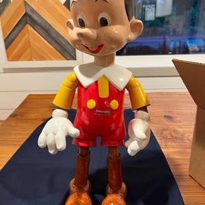 May include: A wooden Pinocchio doll. It features a red outfit, yellow sleeves, and white gloves. The doll has black hair, a white collar, and brown wooden legs and feet. The word "Pinocchio" is printed on the chest.