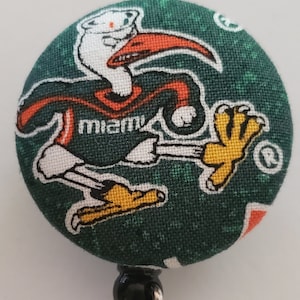 May include: A green and orange fabric button with a cartoon image of a white bird wearing a jersey that says "miami". The button is attached to a black and silver retractable badge holder.