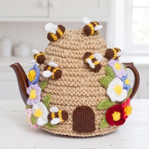 May include: A beige knitted tea cozy shaped like a beehive, featuring several small crocheted bees with brown and yellow stripes and white wings, decorated with colorful crocheted flowers in shades of red, white, blue, and yellow.