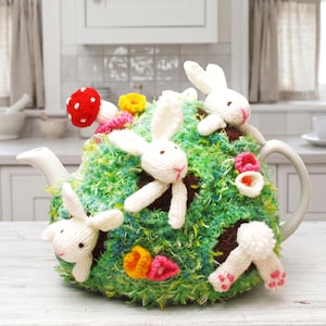 May include: A white ceramic teapot covered by a green textured crochet cozy featuring three white rabbits, a red mushroom with white spots, and small yellow and pink flowers. One rabbit is shown from the back with a white pom-pom tail.
