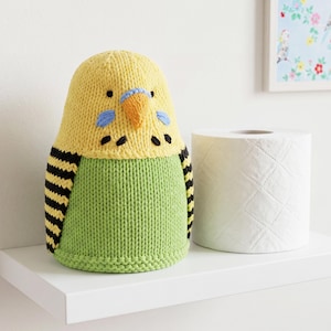 May include: A knitted budgie-shaped toilet paper cover featuring a yellow head with blue cheek patches, a small orange beak, and black eye details. The body is light green with black and yellow striped wings on the sides, crafted in a textured knit.