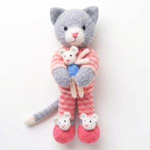 May include: A hand-knitted grey cat plush toy wearing pink and white striped pyjamas. The cat holds a small white mouse toy with a blue shirt. The cat wears white mouse-shaped slippers on its feet and has a long grey tail extending to the left.