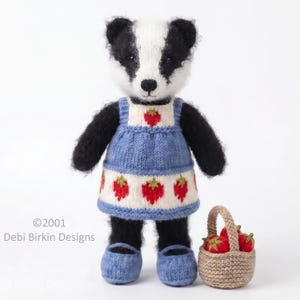 Pode incluir: Um brinquedo de texugo tricotado com pelo preto e branco, vestindo um vestido azul e creme com padrões de morangos vermelhos e sapatos azuis a condizer. Ao lado do texugo está um pequeno cesto entrançado castanho com três morangos.