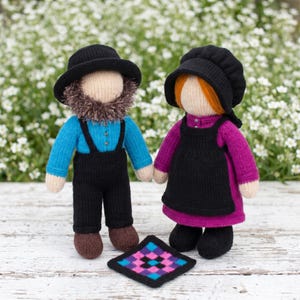 May include: Two knitted dolls, one with a beard wearing a black hat, blue shirt, and black overalls, and one with orange hair wearing a black bonnet, magenta dress, and black apron, with a small square quilt patch placed in front of them.