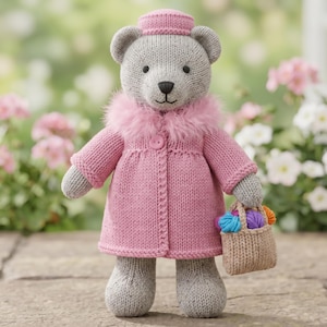 May include: A knitted grey teddy bear wearing a pink coat with a faux fur collar and a matching pink pillbox hat. The bear holds a small tan knitted basket filled with several colourful balls of yarn in its right hand, standing upright.