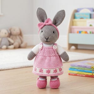 Puede incluir: Un juguete de conejo de peluche tejido con pelaje gris, que lleva un vestido rosa con un dobladillo blanco con un patrón floral rosa, una diadema rosa a juego con un lazo y zapatos rosas. El conejo tiene mangas blancas y dos botones blancos.