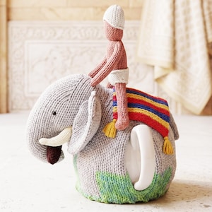 May include: A grey knitted tea cozy shaped like an elephant with white tusks and a dark brown trunk tip. A small pink knitted figure wearing a white hat and loincloth sits on the elephant's back, which is covered by a striped red, blue, and yellow blanket.