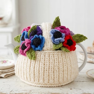 May include: A cream-colored, ribbed knit tea cozy covering a white teapot. The top of the cozy is decorated with a cluster of crocheted flowers in shades of red, blue, purple, and pink, accented with green crocheted leaves and a central handle.