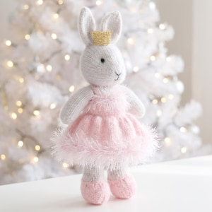 May include: A knitted rabbit doll with light gray fur, wearing a pink fuzzy dress and a small gold glitter crown on its head. The rabbit has black bead eyes and a stitched nose, with matching pink knitted shoes on its feet.