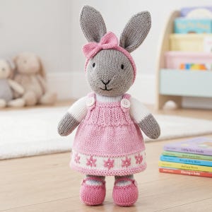 May include: A knitted stuffed rabbit toy with grey fur, wearing a pink dress with white floral embroidery, a matching pink headband with a bow, and pink shoes. The rabbit has white sleeves, two white buttons on the dress straps, and black eyes.