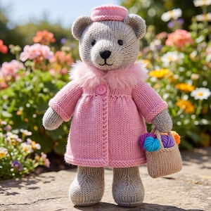 Op de afbeelding: Een grijze gebreide teddybeer met een roze gebreide jas met een kraag van imitatiebont, een bijpassende roze muts en een enkele roze knoop. De beer houdt een klein beige gebreid mandje vast met daarin verschillende kleine bolletjes gekleurde wol.
