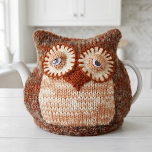 Peut inclure: Une housse de théière tricotée en forme de hibou, dans des tons marron et crème, avec deux grands yeux circulaires en laine blanche et marron, des yeux mobiles en plastique et un bec pointu marron, recouvrant une théière en céramique blanche.