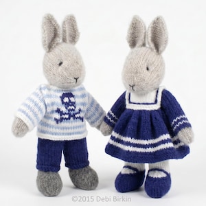 May include: Two knitted rabbit dolls with light grey yarn bodies. The left rabbit wears a light blue and white striped jumper with a dark blue skull and crossbones design and dark blue trousers. The right rabbit wears a dark blue dress.