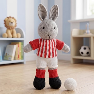 Puede incluir: Un peluche de conejo tejido en color gris que viste una camiseta a rayas rojas y blancas, pantalones cortos blancos, calcetines rojos y zapatos negros. El conejo tiene orejas largas, ojos bordados negros y una nariz pequeña. Una pelota blanca tejida.
