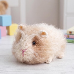 May include: A small, fuzzy, cream and light brown knitted guinea pig toy with a dark brown eye and a stitched pink nose. The plush animal features a soft, textured yarn finish and rounded ears, crafted with detailed needlework to mimic a rodent.