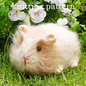 May include: A fuzzy, cream and light brown knitted guinea pig toy with a small pink stitched nose and dark eyes. The text "knitting pattern" is visible at the top of the image. The toy is positioned among green grass and white flowers.