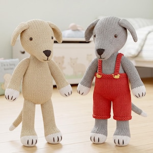 Op de afbeelding: Twee gebreide hondenknuffels. De ene is beige met witte pootjes en een donkerbruine neus. De andere is grijs met witte pootjes, een donkerbruine neus en draagt een rode gebreide tuinbroek met twee kleine gele eendvormige knoopjes aan de voorzijde.