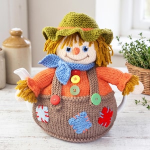 May include: A knitted scarecrow tea cozy featuring a green hat, orange sweater, and brown overalls with colorful patches. The figure has yellow yarn hair, a blue neck scarf, and four buttons on the front. The design includes a stitched face.