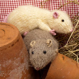 Könnte beinhalten: Zwei weiche, handgestrickte Plüschratten, eine cremefarben und eine hellbraun, mit kleinen schwarzen Perlenaugen, rosa und beigefarbenen gestickten Ohren sowie feinen Garnschnurrhaaren. Die cremefarbene Ratte liegt auf der braunen, beide aus strukturiertem Garn.