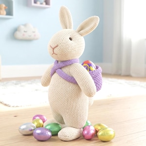 Könnte beinhalten: Ein cremefarbenes Strick-Hasenspielzeug mit langen Ohren und gestickter Nase, das einen violetten Strickschal und einen passenden violetten Rucksack trägt. Der Hase ist von mehreren bunten, glänzenden, in Folie gewickelten Schokoeiern umgeben.